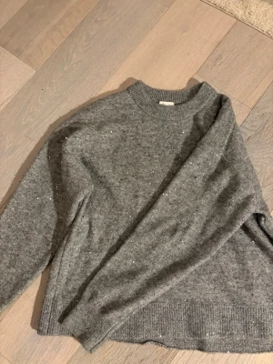 Grå glittrig stickad tröja H&M - Mysig grå stickad tröja från H&M med glittriga paljetter över hela ytan. Rund halsringning och långa ärmar med ribbade mubbar. Materialet är mjukt och värmande, passar bra till kyliga dagar.