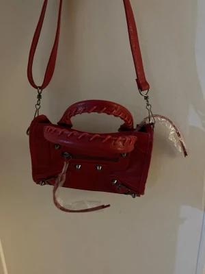Röd handväska i skinn - Cool röd handväska i skinn med snygga metalldetaljer och dragkedja framtill. Väskan har både handtag med flätad look och en avtagbar axelrem. Insidan har spegel och flera fack, perfekt för dina essentials. Statement piece som lyfter din outfit. OBS!! Väskan har inget märke!