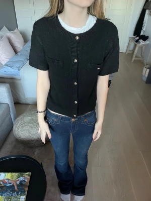 Svart stickad kofta med guldfärgade knappar - Snygg svart stickad kofta från Gina tricot med korta ärmar och guldfärgade knappar framtill. Koftan har två små fickor på bröstet och ribbade detaljer vid hals, ärmslut och nederkant. Perfekt att styla med jeans eller kjol för en cool look. Har tappat ena knappen men går enkelt att fixa till ! 