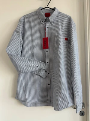 Oanvänd skjorta från Hugo Boss XL - HUGO skjorta – Slim Fit, XL Ny & oanvänd, prislapp kvar.  Blå/vitrandig skjorta i slim fit. Upplevs som något mindre i storleken – se mått.  Mått: • Axel: 49 cm • Bröst (armhåla–armhåla): 56 cm • Ärmlängd: 67 cm • Överarm: 19 cm  Passar dig som normalt har L–XL och vill ha en smalare passform. Perfekt till jobb eller mer uppklädda tillfällen.  Nypris 1299kr