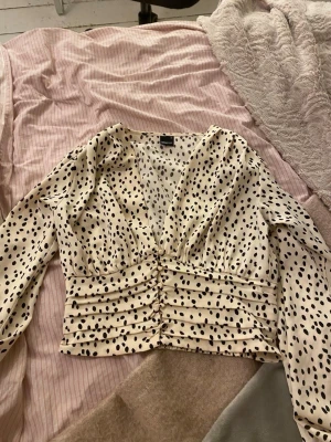 Prickig omlottblus med v-ringning - Säljer en beige omlottblus från Gina  med svarta prickar, djup v-ringning och långärmade puffärmar. Blusen har smockad midja och är croppad.