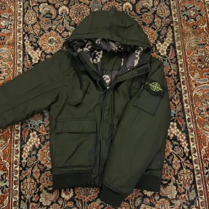Mörkgrön/svart pufferjacka från Stone Island - Säljer en mörkgrön pufferjacka från Stone Island med huva och ribbade muddar. Jackan har flera fickor och en ikonisk patch på ärmen. Insidan är fodrad med ett grått och beige camouflage-mönster. Perfekt för dig som gillar streetwear och vill sticka ut. Storlek large men passar mer som medium