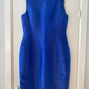 Kornblå ärmlös klänning från Hobbs - Supersnygg kornblå klänning från Hobbs med figurnära passform och klassisk rund halsringning. Klänningen är ärmlös och har en slits baktill för extra rörelsefrihet. Materialet är slätt och har en lätt glansig finish som ger en lyxig känsla. Mått Över bröstet: ca 49 cm Över höften: ca 53 cm Längd från axeln och ner: ca 99 cm  54% ull och 46% silke  Kommer från ett djur och rökfritt hem.