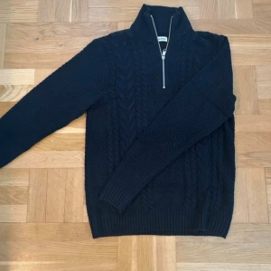 Mörkblå stickad tröja med half zip - Snygg mörkblå stickad tröja från Ralph Lauren med kabelstickat mönster och hög krage. Tröjan har en praktisk half zip-dragkedja och långa ärmar. Perfekt för kyliga dagar och ger en klassisk vibe till din outfit.