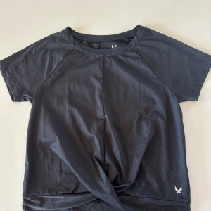 Svart tränings-tshirt från SOC - Svart t-shirt från SOC i funktionsmaterial, perfekt för träning. Modellen har korta ärmar, rund hals och en snygg twistad detalj framtill. Materialet är lätt och andas, vilket håller dig torr och bekväm under aktivitet.