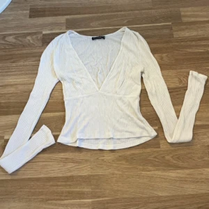 Vit ribbad topp från Brandy Melville - Supersnygg vit ribbad topp från Brandy Melville med djup v-ringning och långa ärmar. Toppen har en figurnära passform och är tillverkad i mjuk bomullsmix. Perfekt att styla med jeans eller kjol för en clean look.