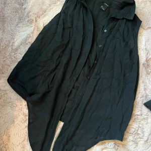 Svart ärmlös knytskjorta - Svart ärmlös knytskjorta från Lindex med klassisk krage och knäppning framtill. Skjortan har två bröstfickor och är tillverkad i ett lätt och luftigt tyg, perfekt för varmare dagar. Snygg att styla lager på lager eller som den är.