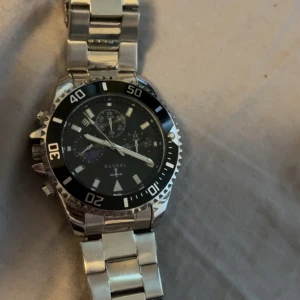 Silverfärgad klocka från TRSOYE - Stilren klocka från TRSOYE med silverfärgad länkarmband och svart urtavla. Klockan har kronograf-funktion, datumvisning och markerade siffror på boetten. Metallarmbandet ger en klassisk och cool vibe, perfekt för dig som gillar detaljer och sportig design.