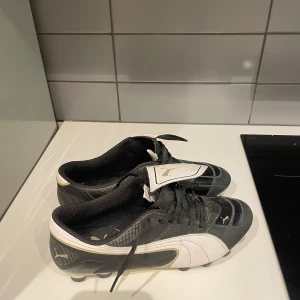 Svarta och vita fotbollsskor från Puma - Säljer ett par klassiska fotbollsskor från Puma i svart och vitt med guldiga detaljer. Skorna har snörning, rund tå och dobbar för bra grepp på planen. Ikonisk Puma-logga på sidan och tungan. Perfekta för dig som vill sticka ut på matchen!