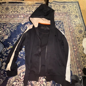 Svart hoodie från Moose Knuckles M - Svart hoodie från Moose Knuckles med beigea detaljer längs ärmarna och insidan av huvan. Tröjan har dragkedja framtill, huva med tryck och långa ärmar. Perfekt för en chill och sportig look. Materialet är mjukt och skönt, troligtvis bomull eller polyester.