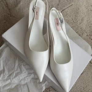 Vita pumps med spetsig tå från Nelly - Stilrena vita pumps från Nelly med spetsig tå och låg klack. Skorna har en öppen häl med justerbart spänne och är tillverkade i syntetmaterial. Perfekta för dig som vill ha en clean och elegant look.