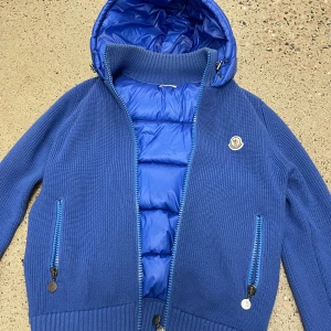 Blå Moncler pufferjacka med huva - Snygg blå pufferjacka från Moncler med stickade ärmar och quiltad front. Jackan har huva, två dragkedjefickor och Moncler-logga på bröstet. Perfekt för dig som vill ha en varm och stilren jacka med sportig känsla.