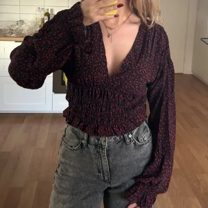 Svart blommig omlottblus med v-ringning - Supersnygg svart blus med små röda blommor, djup v-ringning och omlottkänsla. Har satt några stygn i bysten för att v ringningen var för djup, men det går att ta bort!! Den har tyvärr inte kommit till någon användning💓🫶🏻