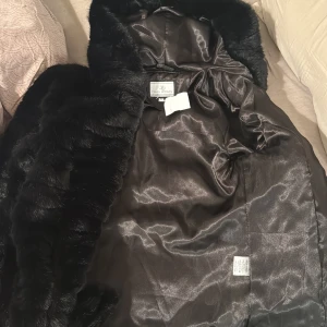 Svart pälsjacka med huva från Flash Woman - Mysig svart pälsjacka från Flash Woman med huva och glansigt innerfoder. Jackan har dragkedja framtill och är helsvart med en fluffig känsla. Perfekt för kalla dagar när du vill vara både varm och snygg. Helt oanvänd . Köpt för 1000