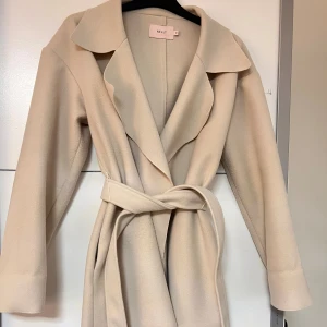 Creme färgad kappa med bälte  - Creme färgad kappa från Nelly med knytskärp i midjan. Fint skick, bara lite knottrig på vissa ställen.