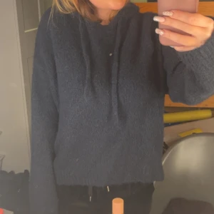 Mörkblå stickad hoodie med luva - Mysig mörkblå stickad hoodie med huva och snörning. Tröjan har lång ärm och en relaxed passform, perfekt för kyliga dagar. Enkel och stilren design som funkar till det mesta.