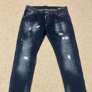 Dsquared2 blå jeans med färgstänk - Snygga blå jeans från Dsquared2 med slitningar, hål och coola färgstänk i rött, gult och vitt. Jeansen har klassisk femficksmodell och raka ben. Tillverkade i Italien i mjukt denimtyg som sitter skönt och ger en trendig look.