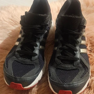 Marinblåa Adidas sneakers med silvriga ränder - Adidas sneakers i marinblå mesh och syntet med silvriga klassiska tre ränder på sidorna. Skorna har snörning, vit och röd platt sula samt rund tå. Perfekta för dig som gillar sportig stil och vill sticka ut med detaljer i silver.