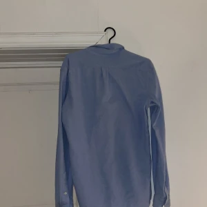 Blå slim fit skjorta från Ralph Lauren - Säljer en klassisk blå skjorta från Ralph Lauren i slim fit-modell. Skjortan är i bomull, har långa ärmar, knappar framtill och ikoniska broderade loggan på bröstet. Perfekt till jeans eller chinos för en clean look.