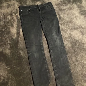 Svarta jeans från Nudie Jeans, W28 L32 - Säljer ett par svarta jeans från Nudie Jeans i storlek W28 L32. De har klassisk femficksdesign, raka ben och subtila detaljer med orange brodyr på bakfickan. Jeansen är tillverkade i mjukt denimtyg och har en snygg, mörk finish.