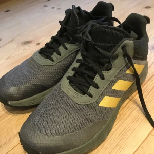Adidas sneakers med gula detaljer - Snygga Adidas sneakers i mörkgrått och svart med gula ränder och logga på sidan. Ovandelen är i mesh och syntet, med svarta snören och robust sula med coolt mönster undertill. Perfekta för dig som gillar sportig stil och vill sticka ut lite extra.