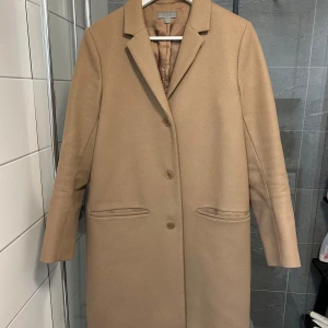 Beige kappa från COS, strl 38 - Snygg beige kappa från COS i klassisk rak modell med krage och tre knappar framtill. Jackan har två fickor och är helfodrad. Perfekt för dig som gillar stilrena och minimalistiska plagg med en modern touch.