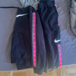 Nike hoodie  byxor med rosa detaljer - Säljer en svart Nike hoodie med stor vit swoosh på bröstet och rosa band med små vita swooshes längs ärmarna. Hoodien har huva och magficka, samt är tillverkad i mjuk bomullsmix. Perfekt för dig som gillar sportig och trendig streetstyle.