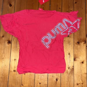 Röd Puma t-shirt med tryck - Röd t-shirt från Puma med stort logotryck i vitt och grått snett över framsidan. Klassisk rund halsringning och korta ärmar. Tillverkad i mjuk bomull, perfekt för en sportig och avslappnad stil.