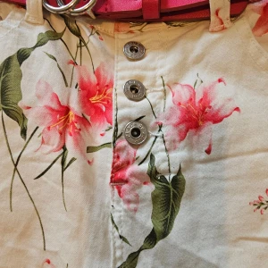 Blommiga croppade byxor med bälte - Snygga croppade byxor med blommigt mönster i rosa och grönt på vit botten. Byxorna har knappar framtill och kommer med ett rosa bälte med silverdetaljer. Perfekt för dig som vill sticka ut med färg och mönster.