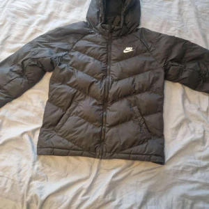 Svart pufferjacka från Nike  - Svart pufferjacka från Nike i storlek XL med huva och dragkedja framtill. Jackan har ett stilrent Nike-logga på bröstet och är tillverkad i syntetmaterial. Perfekt för kalla dagar med sin vadderade design och sportiga look.