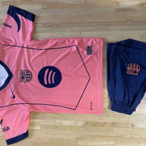 Barça rosa matchtröja Lamine Yamal #10 - Säljer en FC Barcelona fotbollströja i rosa med mörkblå detaljer, tryck och logga. Tröjan har korta ärmar, rund hals och är tillverkad i lätt, ventilerande polyester. Matchtröjan har Lamine Yamal och nummer 10 på ryggen. Shorts i mörkblått med Barça-logga ingår.