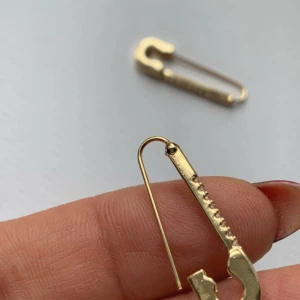 Guldiga örhängen med strass i säkerhetsnålform - Unika örhängen i form av säkerhetsnålar i guldig färg, dekorerade med små glittrande stenar längs ena sidan. Perfekt accessoar för dig som vill sticka ut med en edgy och trendig stil. Säljs som par.