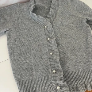 Grå stickad kofta med pärlknappar - Superfin grå stickad kofta med volangkant och pärlknappar framtill. Koftan har långärmade ärmar och ribbade muddar vid ärmslut och nederkant. Knappt använd!