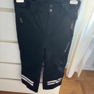 Svarta täckbyxor med reflexdetaljer - Svarta täckbyxor från Lindberg med reflexdetaljer vid bensluten, dragkedjefickor och justerbart bälte i midjan. Perfekta för vintern och utomhusaktiviteter. Funktionell design med robust material och praktiska detaljer.