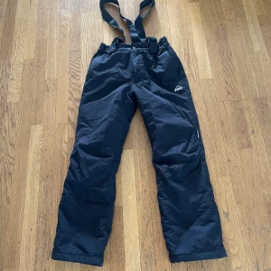 Svarta täckbyxor från McKinley - Svarta täckbyxor från McKinley med justerbara hängslen och elastisk midja. Byxorna har raka ben och är tillverkade i ett slitstarkt syntetmaterial som står emot kyla och vind. Perfekta för vinterns alla äventyr i snön.