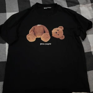 Palm Angels svart t-shirt med björn - Svart t-shirt från Palm Angels med ikoniskt motiv av en isärplockad nallebjörn på framsidan och stor Palm Angels-logga på ryggen. Den är bred i sin klassisk passform och rund hals. Tillverkad i mjuk bomull, perfekt för en streetwear-look.