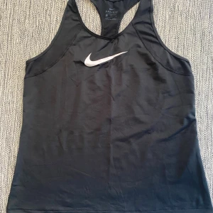 Svart Nike Dri-Fit träningslinne  - Svart sportig linnetopp från Nike med klassisk vit swoosh-logga på bröstet. Modellen har brottarrygg och är tillverkad i Dri-Fit polyester som andas, perfekt för träning. Enkel och stilren design som funkar till alla sportiga outfits.                              Tvättad men aldrig använd  Mått Över bröstet: ca 48 cm Längd från axeln och ner: ca 61 cm  Polyester och elastan  Kommer från ett djur och rökfritt hem.