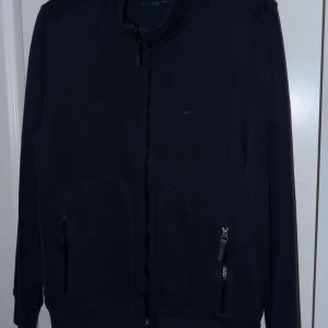 Svart kofta med dragkedja från Henri Lloyd - Svart kofta från Henri Lloyd med hel dragkedja framtill och två fickor med dragkedjor. Långärmad och enkel design, perfekt för lager-på-lager. Tillverkad i mjuk bomull för en skön känsla.