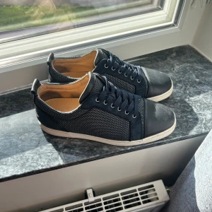 Christian Louboutin mörkblå sneakers - Snygga svarta sneakers från Christian Louboutin med klassisk röd sula och signatur på undersidan. Ovandelen är i mesh och skinn med snörning och rund tå. Insidan är beige och sulan är vit med röd detalj undertill. Perfekt för dig som gillar exklusiva märken och stilren design. Sälj inte längre rare färg självklart äkta.