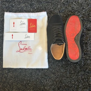 Svarta sneakers från Christian Louboutin - Snygga svarta sneakers från Christian Louboutin i mocka med klassisk röd sula och rund tå. Skorna har svarta snören och en stilren design som passar perfekt till streetwear. Innersulan är beige och de kommer med originalpåse och äkthetskort. Priset är diskuterbart