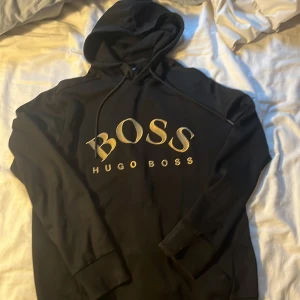 Svart hoodie från Hugo Boss med guldtryck - Svart hoodie från Hugo Boss med stort broderat BOSS-tryck i guld på bröstet. Klassisk huva med snörning och ribbade muddar. Perfekt för dig som vill ha en stilren och lyxig streetwear-look. PRISET GÅR ATT DISKUTERA!!!