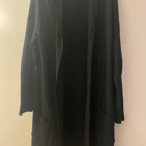 Svart stickad lång cardigan med huva - Svart lång stickad cardigan från bis Missing Link. Jackan har huva, långa ärmar och två stora fickor framtill. Perfekt för lager-på-lager och chill vibes. Enkel och stilren design som funkar till det mesta. ( skärp saknas) stl:M Material: Acryl, Ull samt Viscose. 