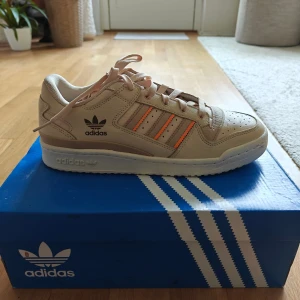 Adidas Forum Low CL beige sneakers - Säljer ett par Adidas Forum Low CL sneakers i beige skinn med klassiska tre ränder i orange och grått på sidorna. Skorna har snörning, perforerad tå och vit platt sula. Perfekt för dig som gillar retrovibbar och stilren design.