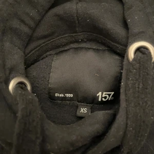 Svart hoodie från 157 med huva XS - Svart hoodie från 157 i storlek XS. Klassisk modell med huva och snörning framtill, samt långa ärmar. Perfekt för en chill och avslappnad stil. Mjuk och skön bomullskänsla.
