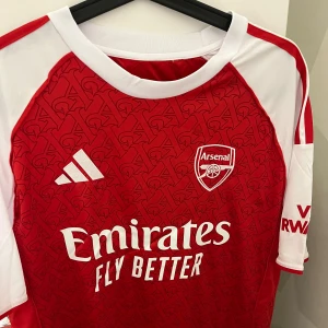 Arsenal hemmatröja Adidas röd/vit - Snygg Arsenal hemmatröja från Adidas med klassiskt röd front och vita ärmar. Tröjan har tryckt klubbmärke, Adidas-logga och sponsortryck. Materialet är lätt och andas, perfekt för match eller träning.  Priset går att diskutera vid snabb affär 
