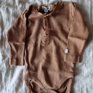 Ribbad brun långärmad body Newbie - Superfin ribbad långärmad body från Newbie i en varm brun nyans. Bodyn har knappdetalj framtill och är tillverkad i mjuk bomull, perfekt för mysiga dagar. Smidig knäppning i grenen för enkel påklädning. Nån liten noppra vid knapparna men inga andra skavanker vad jag har kunnat se.