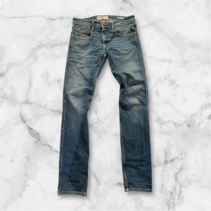 Replay Anbass blå jeans herr 31/32 - Snygga blå Replay Anbass jeans med klassisk femficksdesign och slitna detaljer. Jeansen har raka ben, normal passform och är tillverkade i slitstarkt denim. Märkespatch bak i midjan och diskret Replay-logga vid fickan. Perfekta för en avslappnad streetstil.