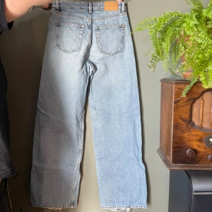 Ljusblå wide jeans från Monki, stl 26 - Säljer ett par ljusblå jeans från Monki i storlek 26. Jeansen har raka, vida ben och klassisk femficksdesign. Tillverkade i bomull med snygg tvättad look och råa kanter nertill. Något slitna i nerkanten av byxbenen, se bild, i övrigt i fint skick.
