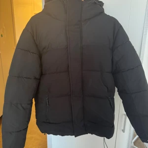 Svart pufferjacka från Zara - Svart pufferjacka från Zara med huva och dragkedja framtill. Jackan har två fickor med dragkedja och en enkel, stilren design. Perfekt för kalla dagar och har en skön, vadderad känsla. Passformen är normal och jackan är helsvart utan mönster.