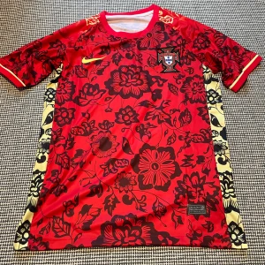 Portugal Nike röd blommig fotbollströja - Snygg Portugal fotbollströja från Nike i rött med svarta blommönster och gula detaljer längs sidorna. Broderat landslagsmärke på bröstet. Tröjan är kortärmad och tillverkad i lätt, ventilerande material som passar perfekt för match eller träning.